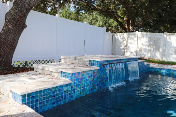 WinWay Homes Custom Pool with Blue Tile Water Feature, White Privacy Fence, Stone Coping & Tropical Backyard Setting