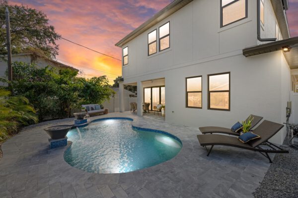 WinWay Homes Rear Exterior at Dusk with Curved Pool, Lounge Chairs, Covered Patio & Illuminated Windows