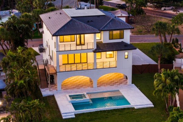 WinWay Homes Elevated Coastal Home at Dusk with Pool, Lit Windows, White Exterior, Covered Entry & 2-Car Garage