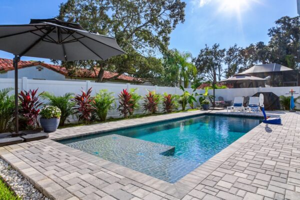 WinWay Homes Backyard Pool with Sun Shelf, White Privacy Fence, Paver Decking & Tropical Plantings