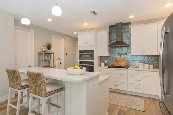 WinWay Homes Kitchen with white peninsula, woven bar stools, decorative range hood, white cabinetry & coastal-inspired finishes